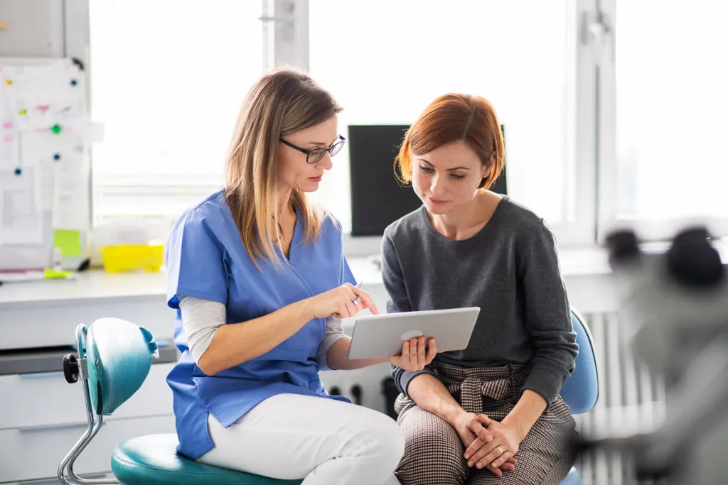 Female dentist using tablet to explain dental treatment plan to attentive female patient in modern dental office.