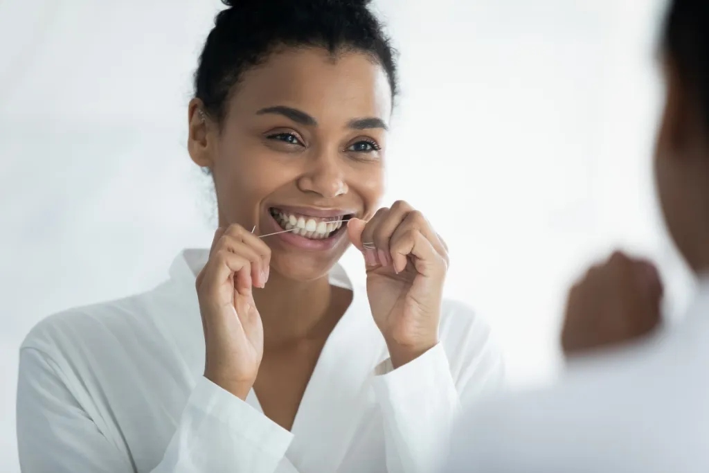 Black woman flossing teeth while looking in the mirror, maintaining oral health.