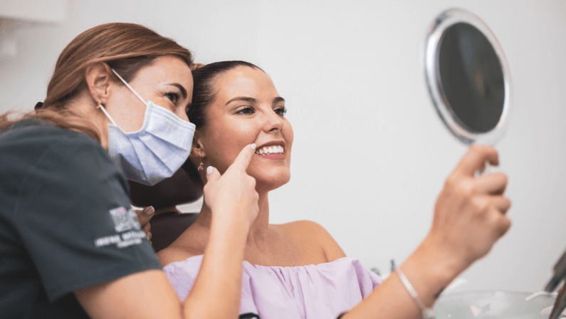 The woman looks at her smile with a mirror in a clinic. Dental clinic. Well-groomed white teeth after being treated by a doctor. the woman dentist points to the smile of the patient.