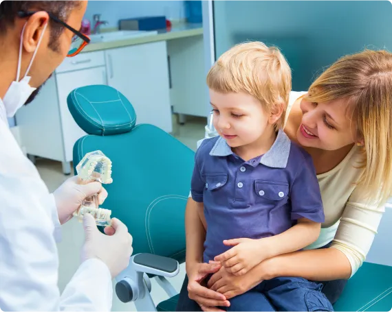 Kind dentist uses a dental model to patiently explain oral care to a young boy with his mother