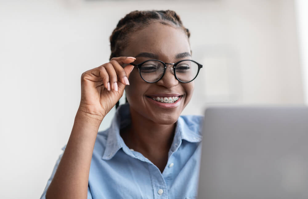 Young Specialist. Smiling african american lady with stylish eyeglasses and braces using laptop at workplace in office, working on computer, looking at screen and smiling, closeup shot, free space