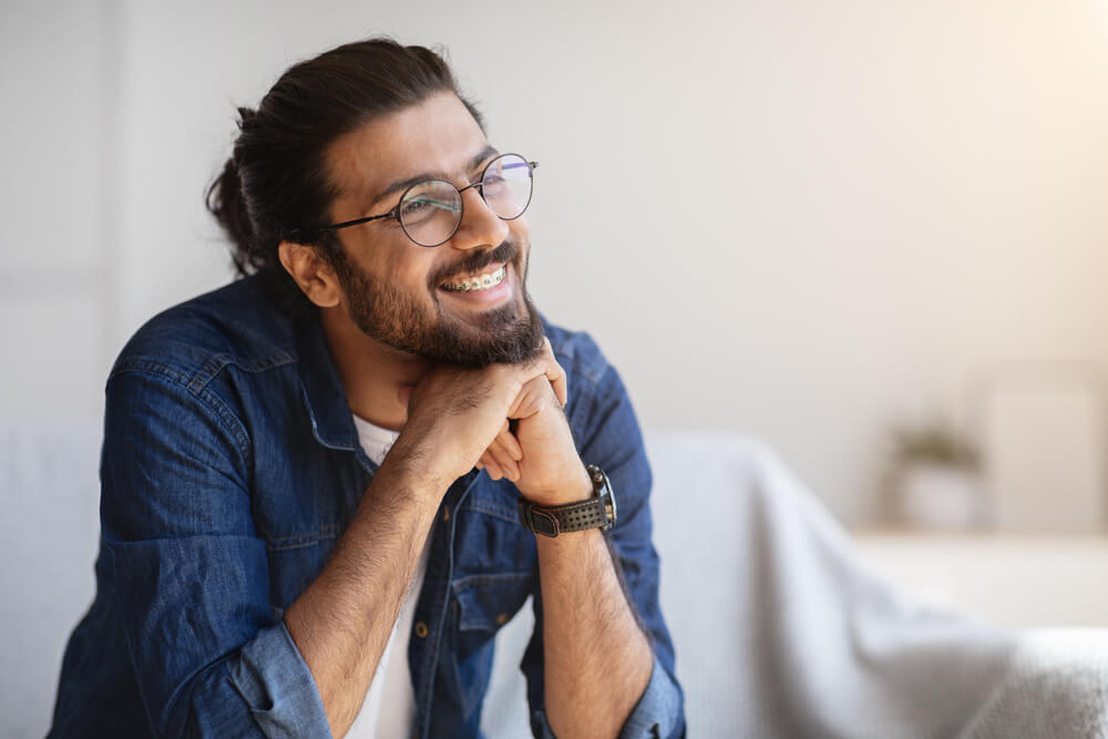 Portrait Of Smiling Indian Man With Eyeglasses And Braces In Home Interior, Handsome Pensive Western Guy Daydreaming And Looking Away, Thinking About Something, Selective Focus With Copy Space
