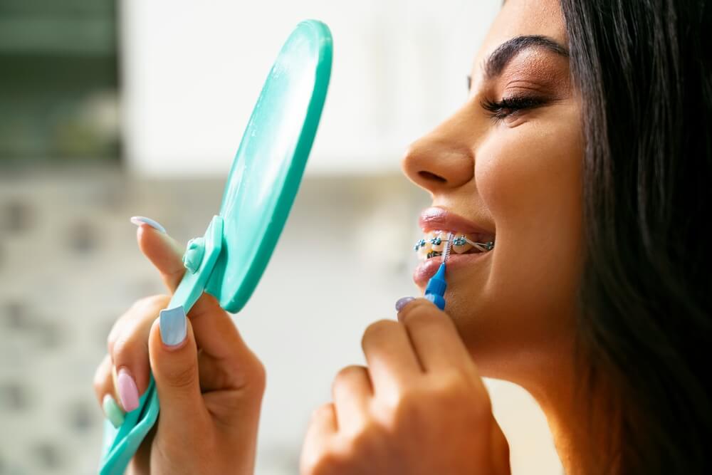 Pretty satisfied young woman holding mirror and interdental brush, dental braces oral hygiene concept