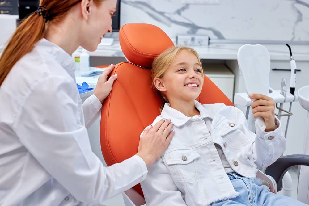 Cute child girl with healthy smile sit looking at mirror during consultation with dentist at dental office, side view. Kid is sitting on couch waiting for treatment, don't afraid of stomatologist