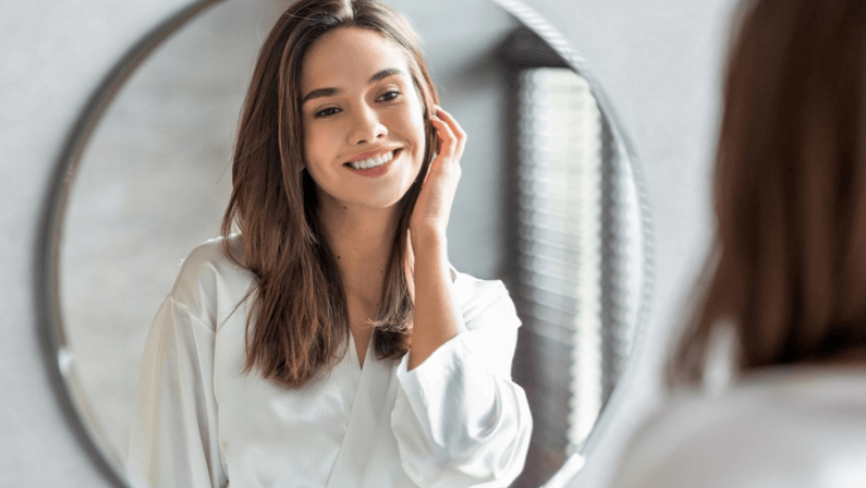 Beauty Concept. Portrait Of Attractive Happy Woman Looking At Mirror In Bathroom, Beautiful Millennial Lady Wearing White Silk Robe Smiling To Reflection