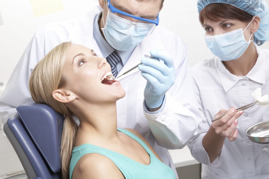 Male dentist examines female patient's teeth with dental hygienist assisting