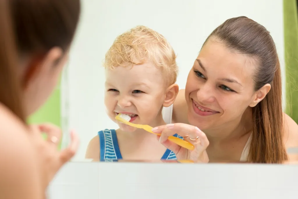 Mom smiles attentively while watching her baby boy brush his teeth, fostering healthy dental hygiene habits from a young age.