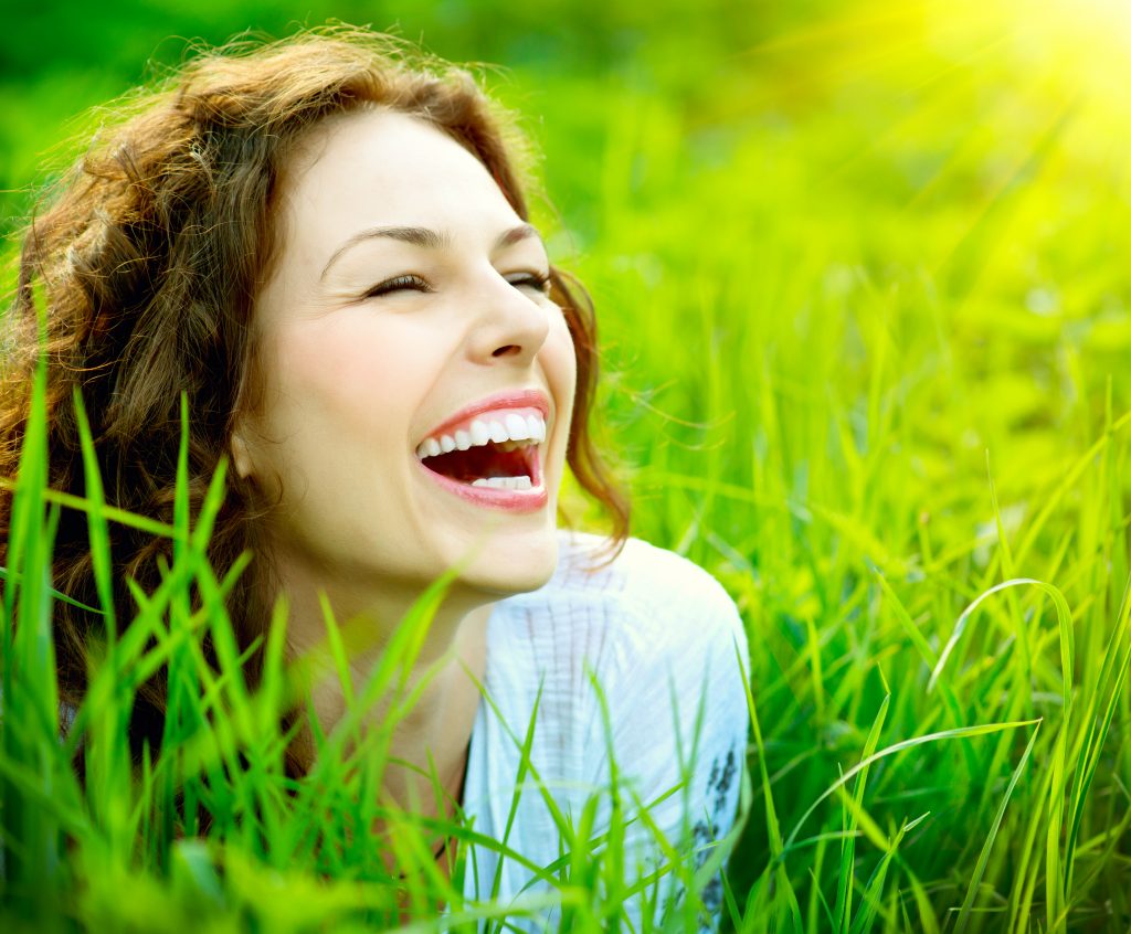 Young woman smiles brightly, revealing healthy teeth, with a grassy background.