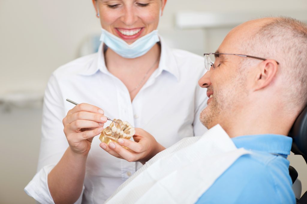 Senior man smiles attentively while listening to female dentist explain dental treatment using a model. Modern dental office.