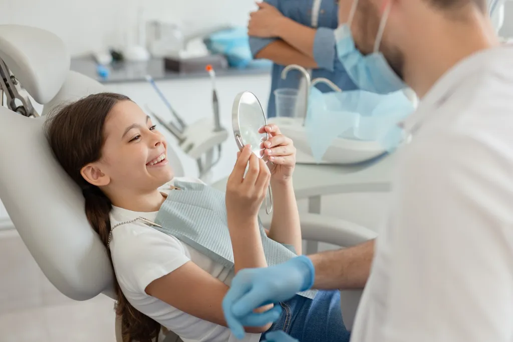 Young woman reclines comfortably in dental chair, attentively examining her teeth in the mirror during dental checkup.