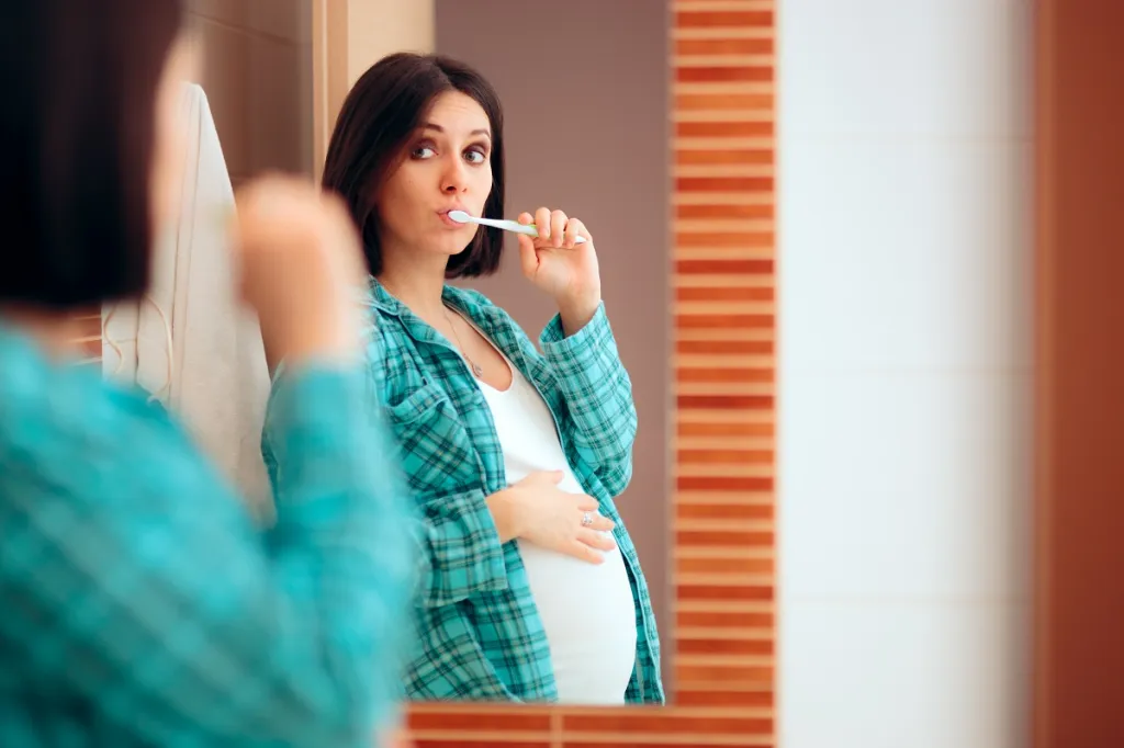 Pregnant woman brushing her teeth diligently while looking in the mirror, maintaining good oral hygiene during pregnancy.