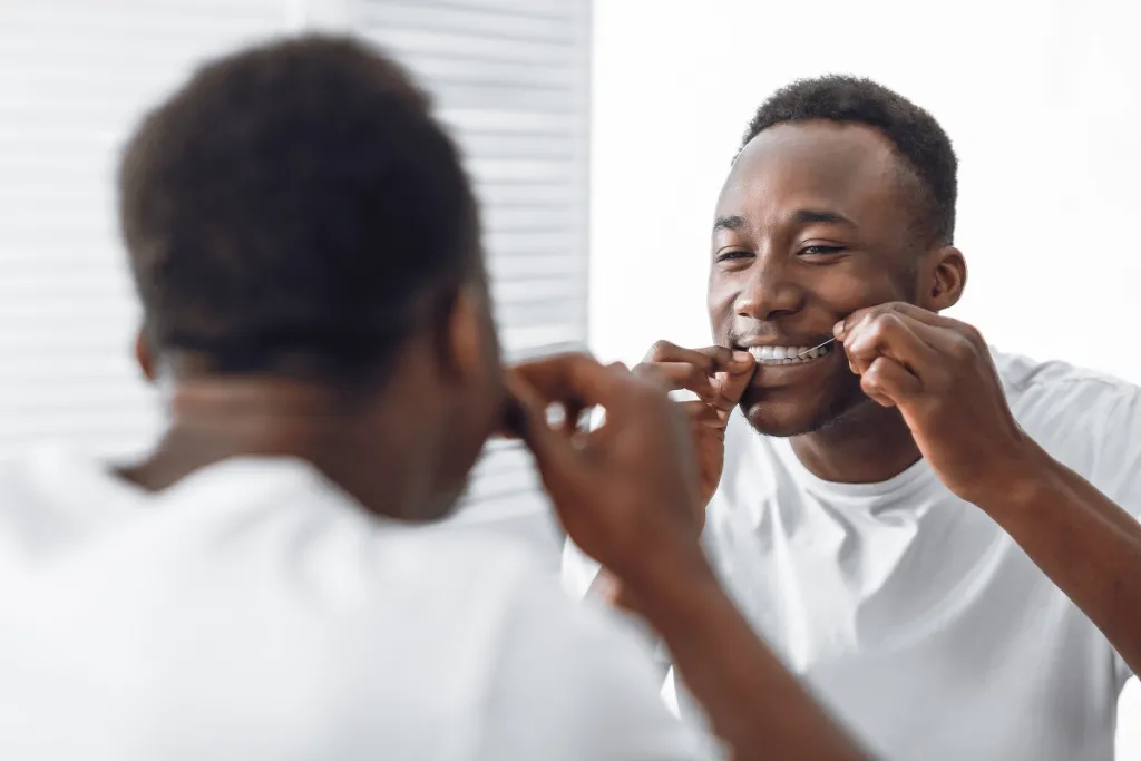 Black man flossing teeth while looking in the mirror, maintaining oral health.