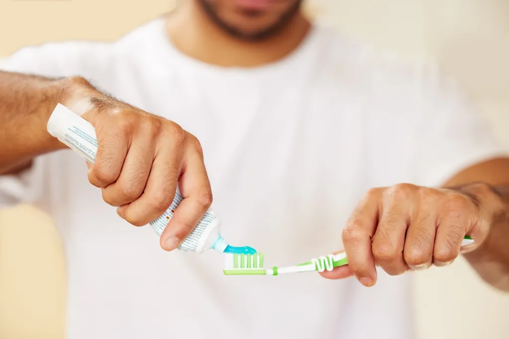 Man applying toothpaste to toothbrush in bathroom, ready for morning oral hygiene routine.