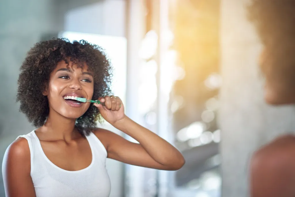 Young Black woman maintaining oral hygiene by brushing teeth in front of bathroom mirror