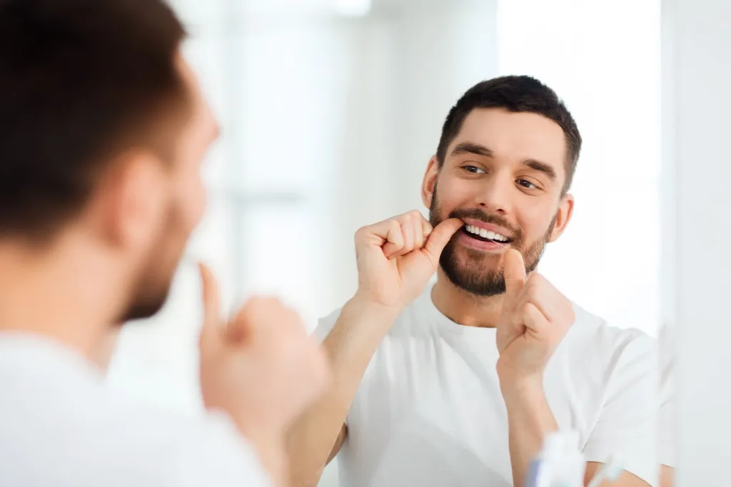 Man maintaining oral hygiene by flossing teeth while looking in bathroom mirror